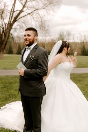 Bride and groom stand back-to-back outdoors, groom in gray suit smiling, bride in white ballgown holding phone.