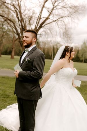 Smiling bride and groom stand back-to-back holding vow books in a green park setting on their wedding day.