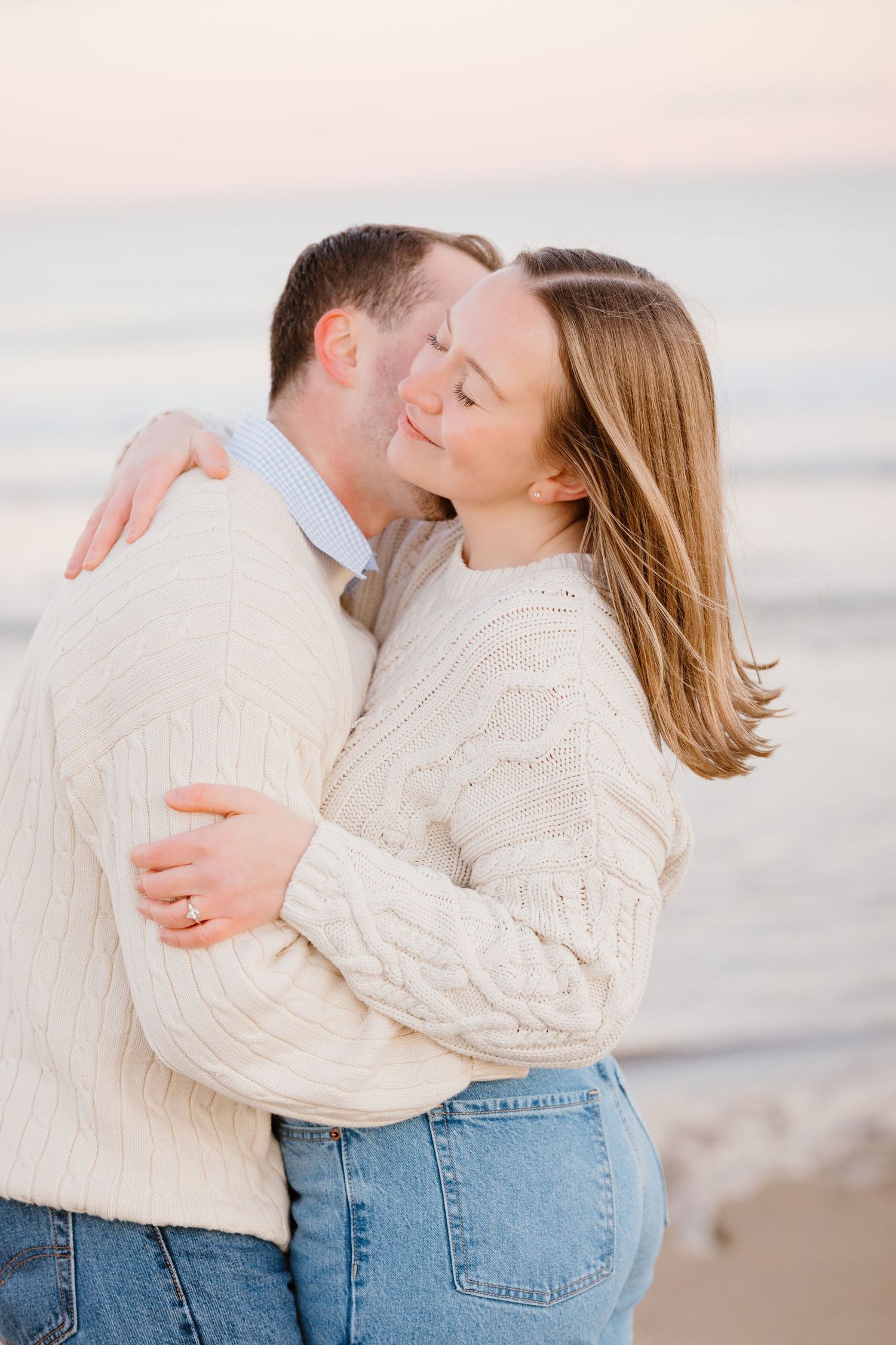 Couple in matching cream cable-knit sweaters and jeans embracing on a beach at sunset.