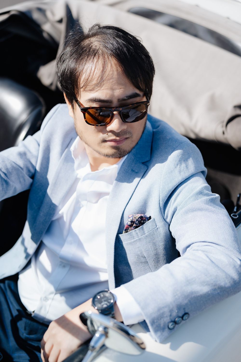 A stylish Asian man in a light blue blazer and sunglasses sits in a convertible car, exuding sophistication.