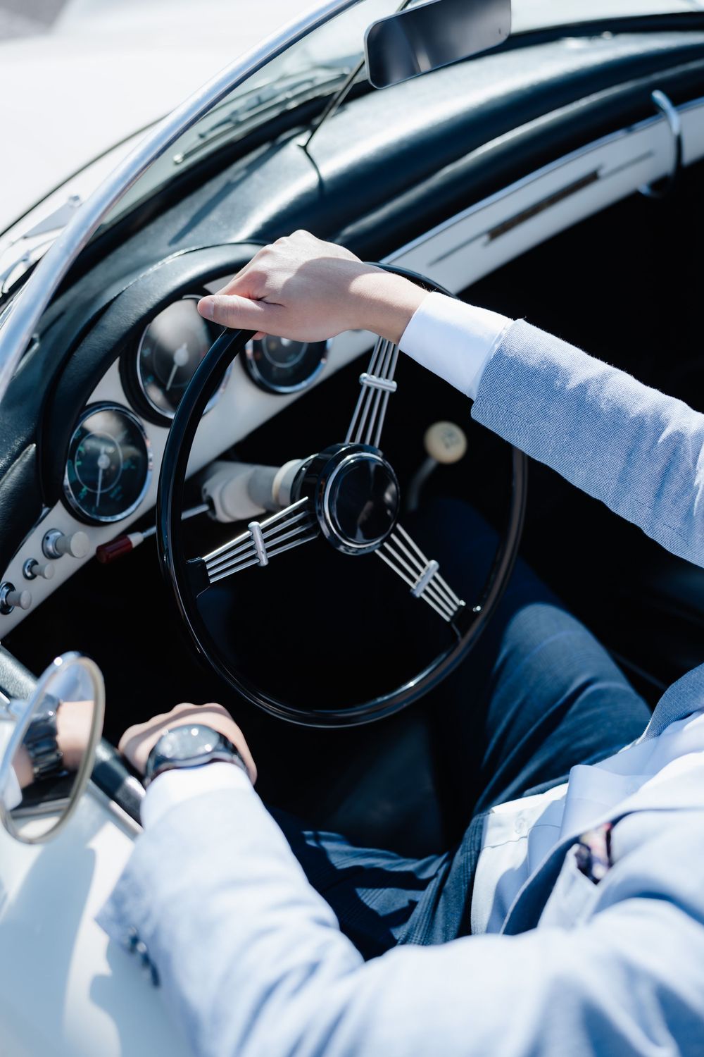 Person in a light blue suit driving a vintage classic car with a retro three-spoke steering wheel.