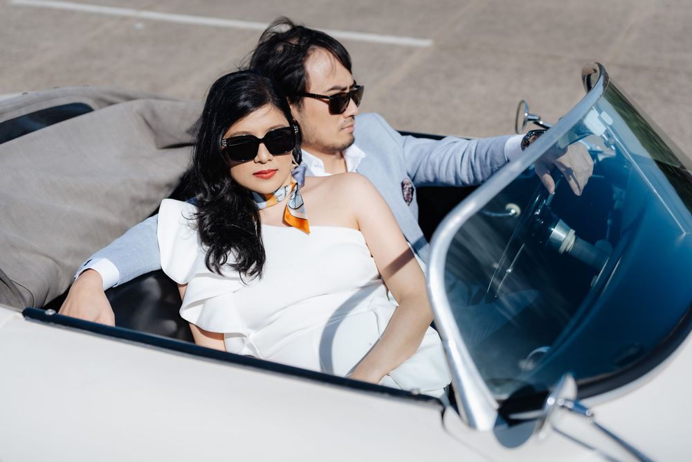 Stylish couple in sunglasses riding in a classic white convertible car, dressed in elegant white outfits.