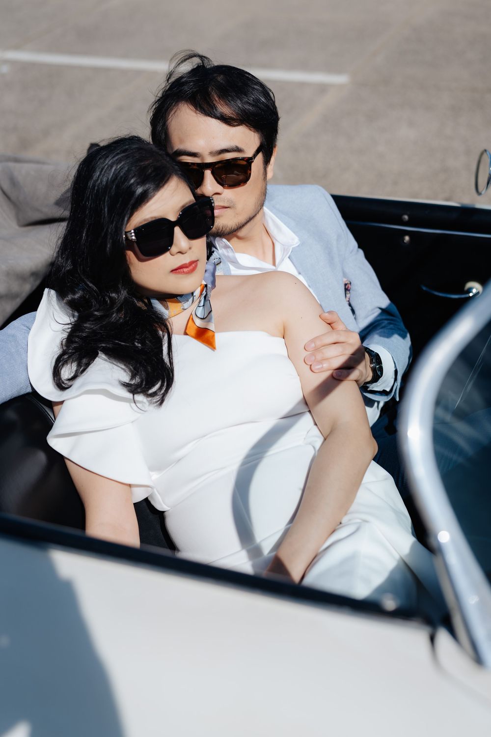 Stylish couple in sunglasses and white outfits sitting closely together in a classic convertible car on a sunny day.