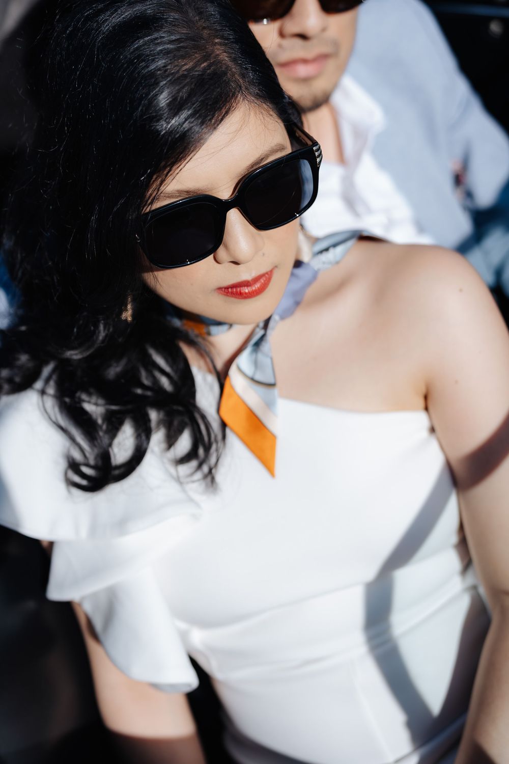 A stylish woman in white off-shoulder top and black sunglasses poses with a man in sunglasses behind her.