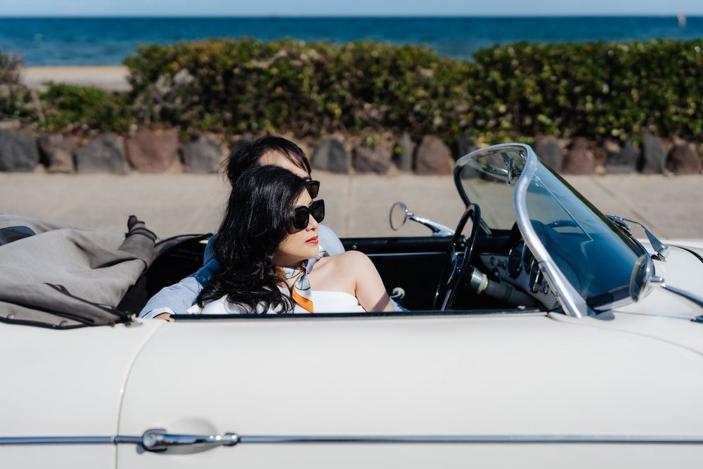 Woman in sunglasses driving a white vintage convertible car near a sunny beach.
