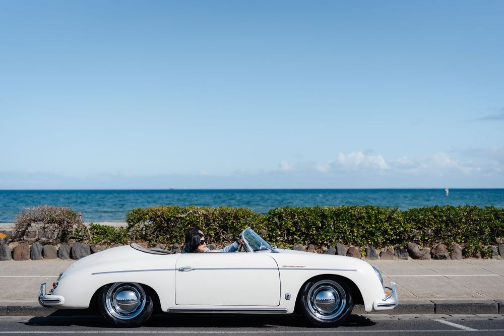 White vintage Porsche 356 convertible parked by the ocean on a sunny day with a clear blue sky.