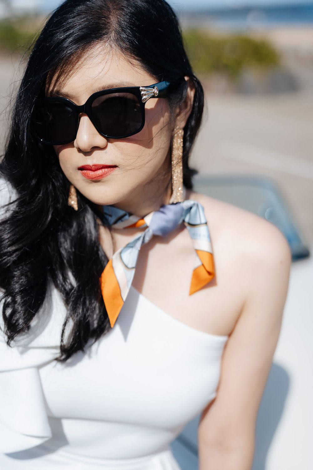 Stylish Asian woman in white dress, black sunglasses, silk scarf, and gold earrings posing outdoors on a sunny day.