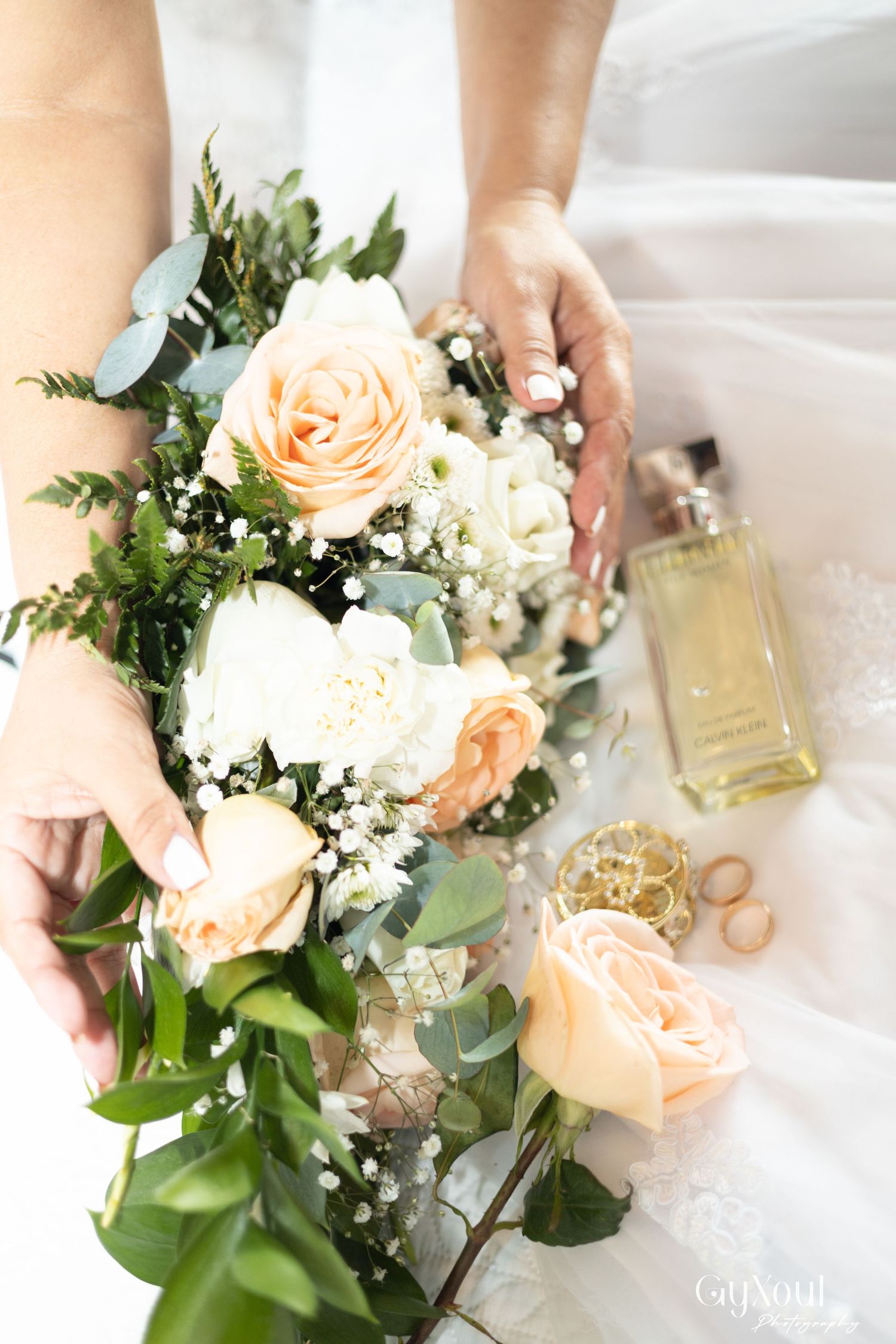 Bride holding a bouquet of peach and white roses with greenery, beside gold rings and perfume bottle.