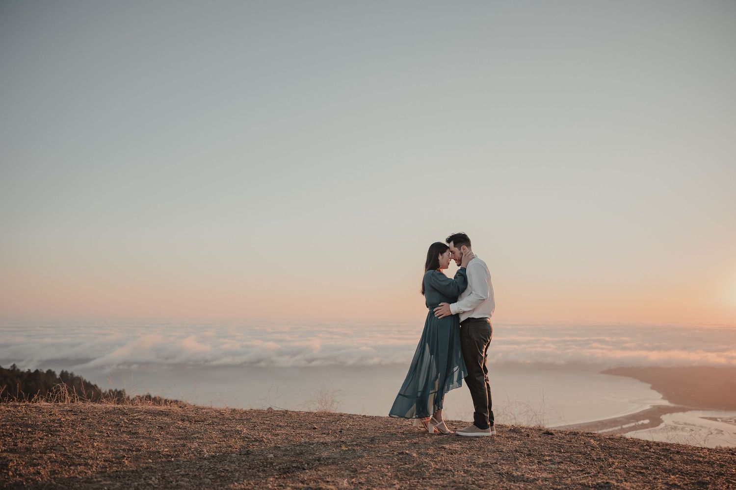 Silhouetted couple embraces on mountain peak overlooking misty coastal sunset view.