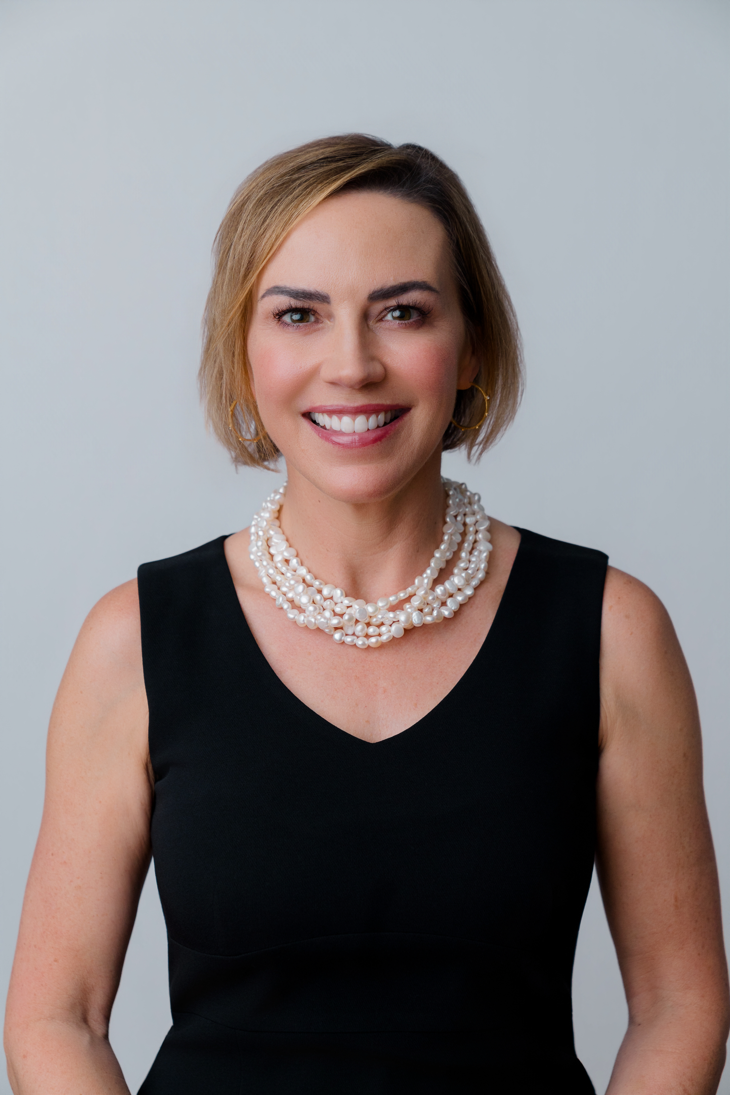 Professional woman in black sleeveless dress and pearl necklace smiling against gray background.