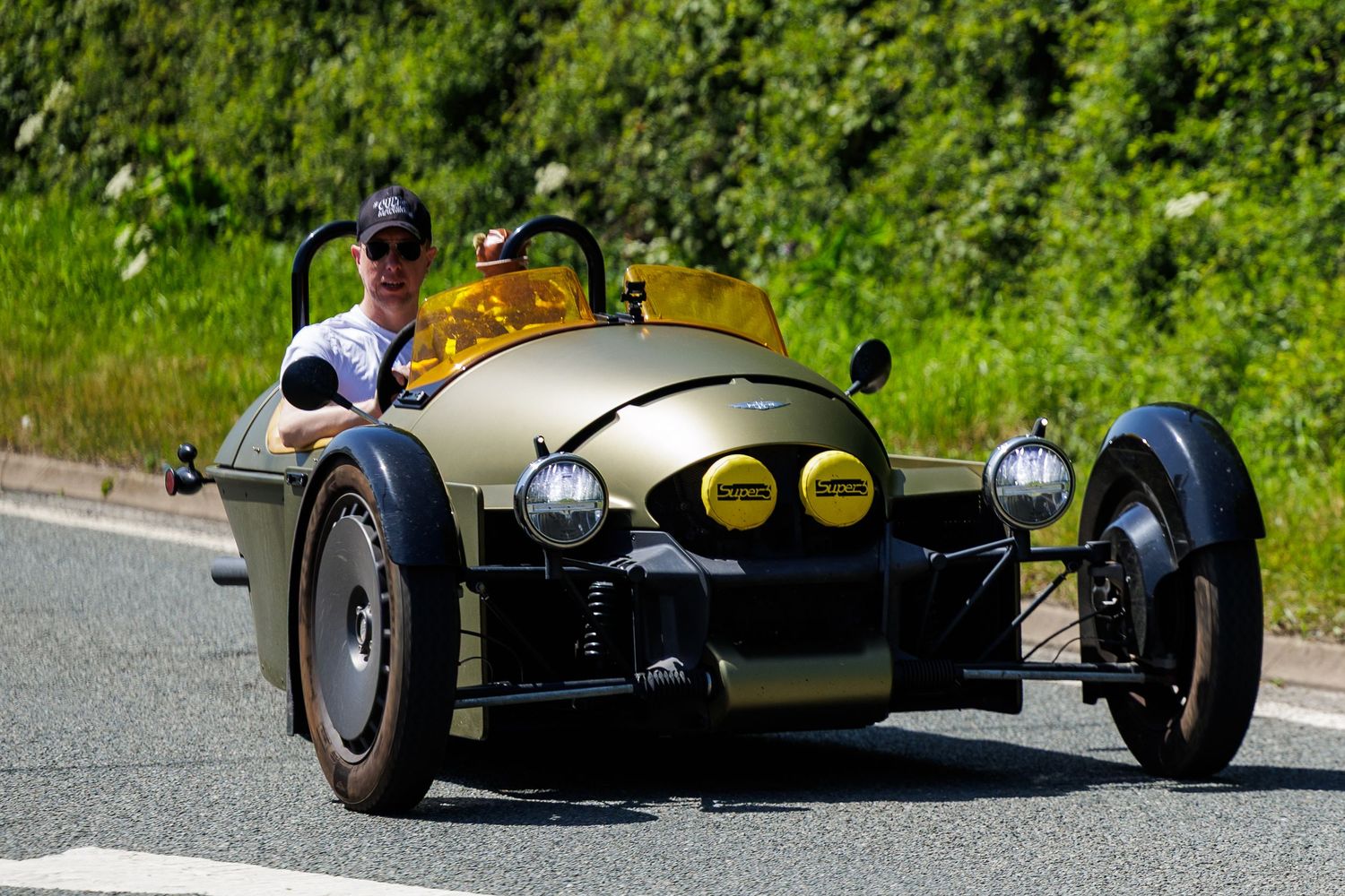 Vintage gold three-wheeled Morgan roadster driven by a smiling man on a sunny road.
