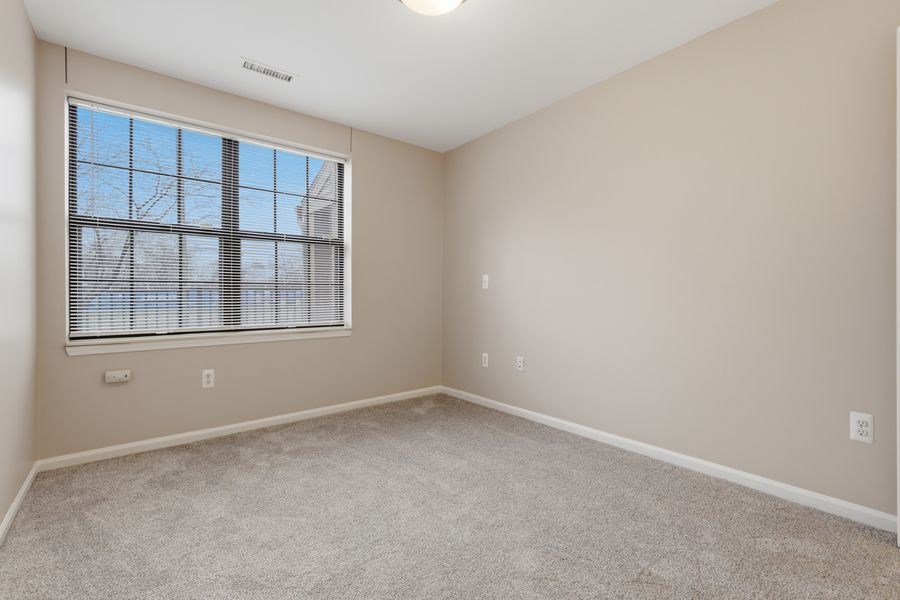 Bright empty bedroom with beige carpet, neutral walls, white baseboards, and a large window with blinds.