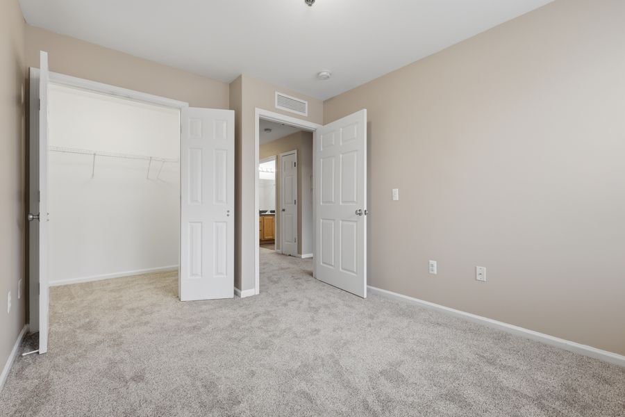 Bright bedroom with neutral carpet, open closet, and white doors leading to hallway in a modern apartment.