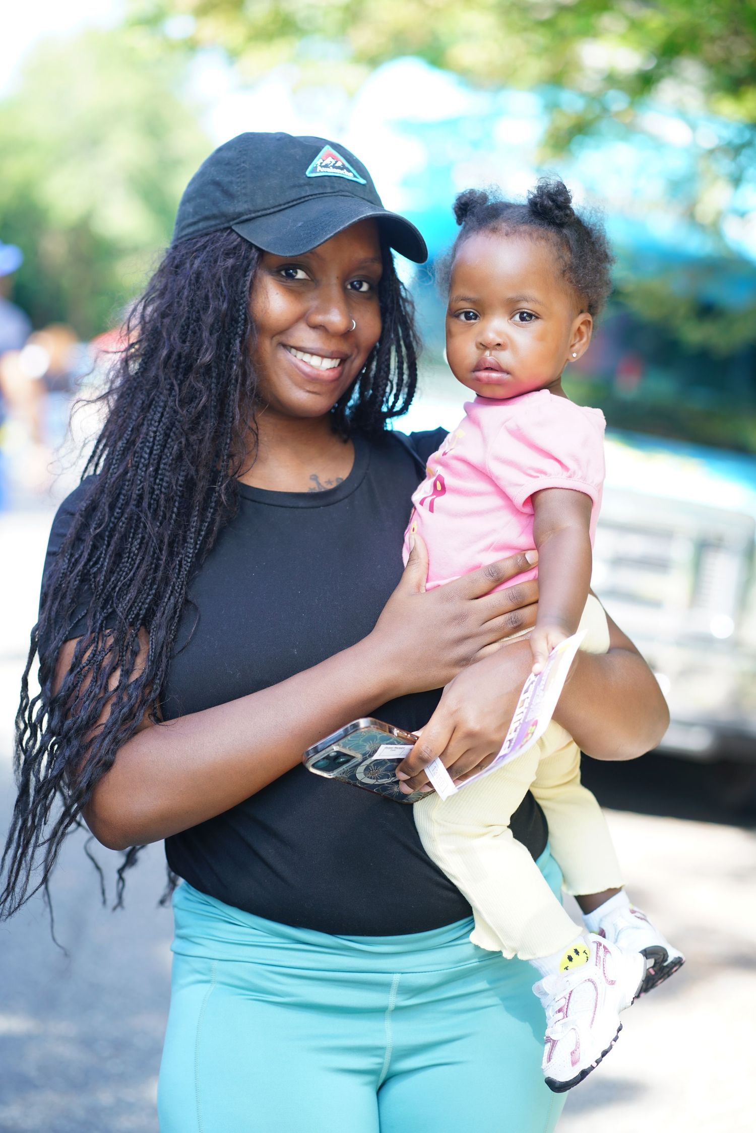 A smiling woman in a black cap holds a young toddler girl in a pink shirt outdoors.
