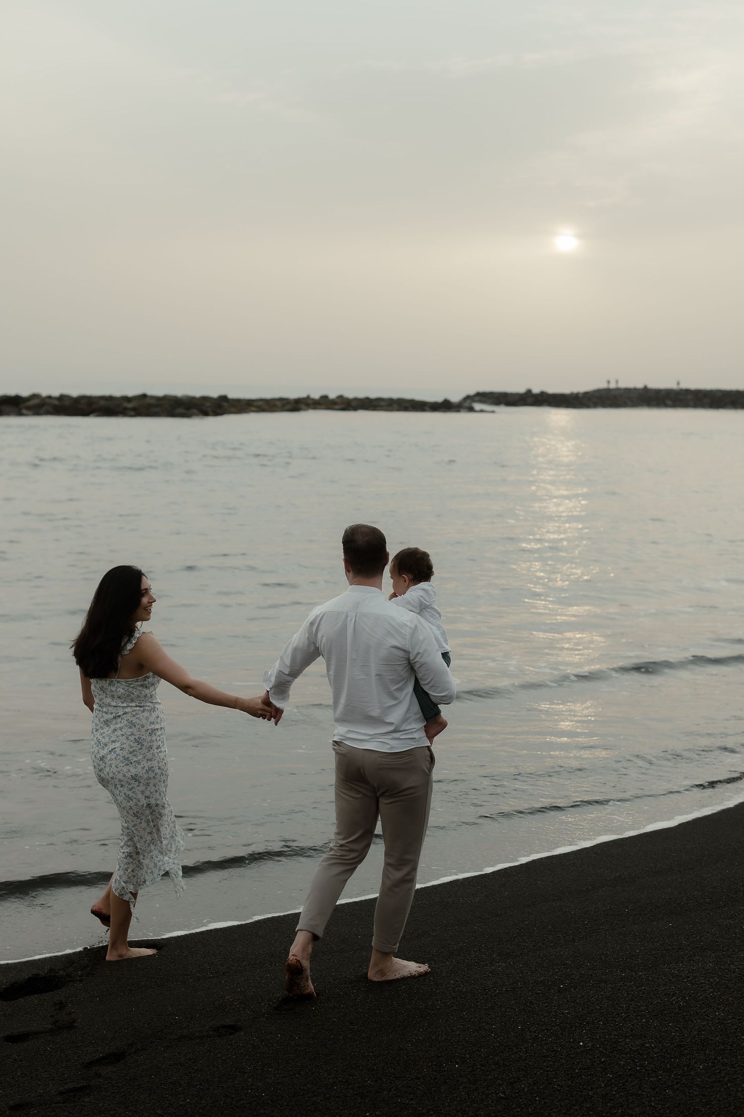 Family of three walking hand-in-hand on a black sand beach at sunset, parents carrying young child.