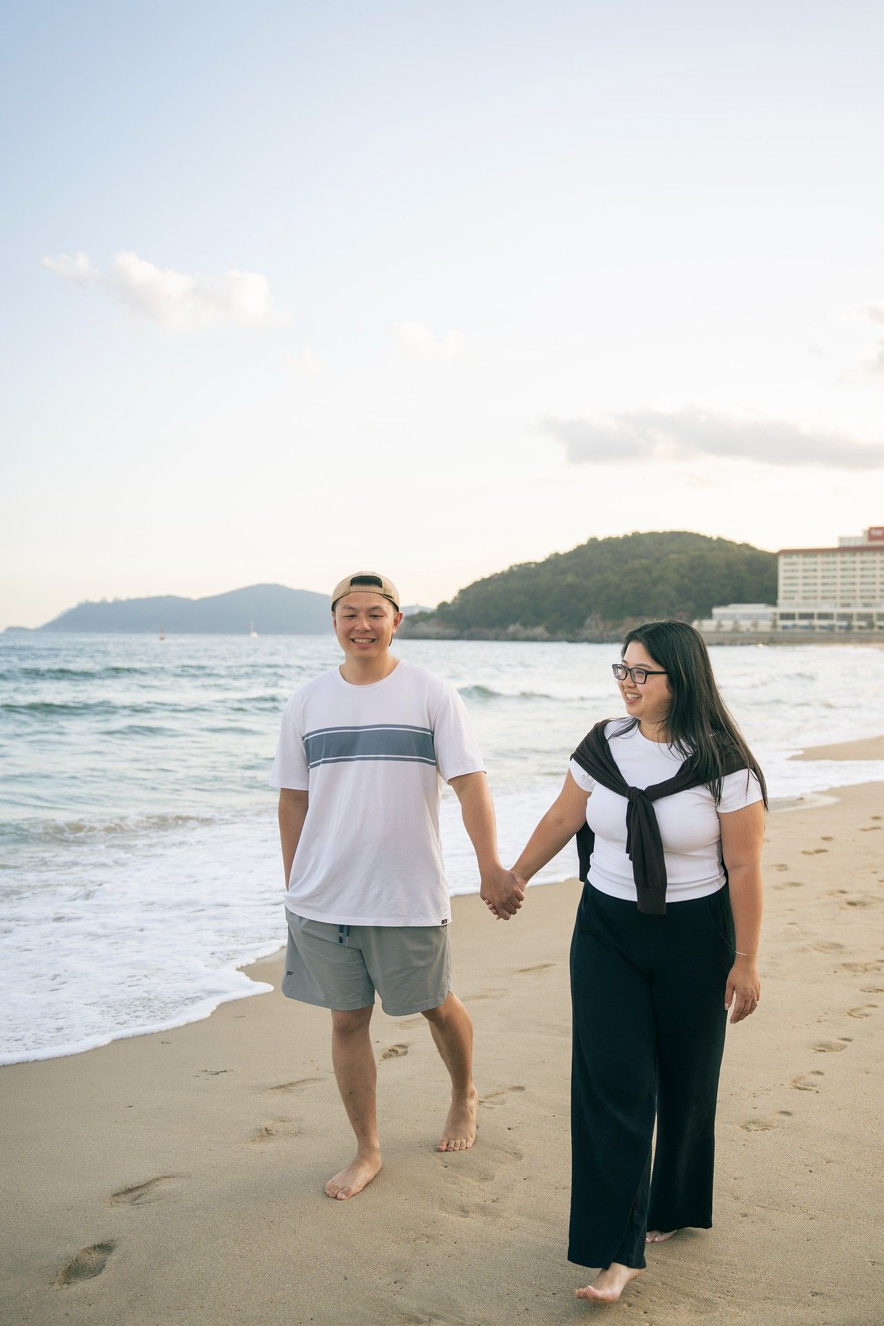 Couple holding hands and walking barefoot on a sandy beach at sunset with ocean waves and green hills in the background.