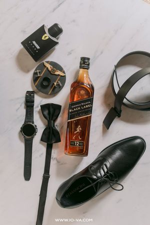 Flat lay of groom essentials featuring Johnnie Walker Black Label whisky, dress shoes, bow tie, watch, belt, and cologne on marble.