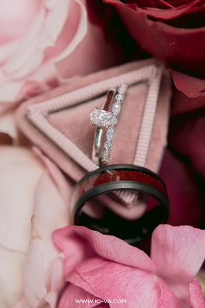 Oval diamond engagement ring with diamond band and black tungsten wedding band resting on pink roses and velvet ring box.