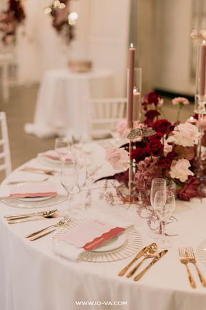 Elegant wedding table setting with gold cutlery, pink menus, crystal glasses, and red and pink floral centerpiece with candles.
