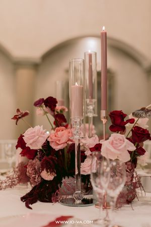 Elegant wedding centerpiece with pink and burgundy roses, tall glass candleholders, and lit taper candles on a white tablecloth.