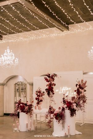 Elegant wedding ceremony backdrop with burgundy floral arrangements, neon sign, candles, and fairy lights in a romantic venue.