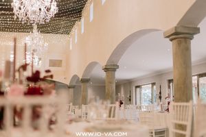 Elegant wedding reception hall with arched columns, crystal chandelier, fairy lights, and romantic red floral centerpieces on white tables.