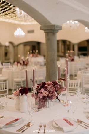 Elegant wedding reception table with burgundy and mauve floral centerpiece, tall taper candles, and white linens in a grand ballroom.