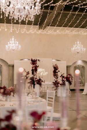 Elegant wedding reception hall with crystal chandeliers, string lights, burgundy floral arrangements, and a neon sign backdrop.