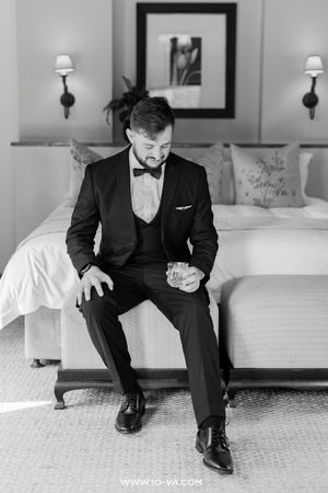 Groom in black tuxedo sitting on bed holding a glass in a classic black and white wedding photo.