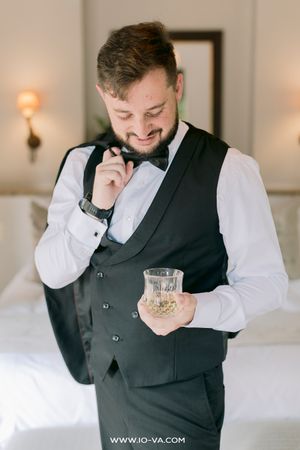 Smiling groom in black vest and bow tie holding a whiskey glass while adjusting his collar in a hotel room.
