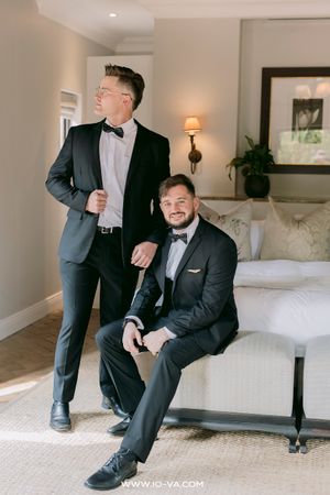 Two grooms in black tuxedos with bow ties pose together in an elegant hotel room before their wedding ceremony.