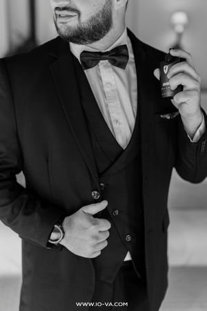 Black and white photo of a groom in a three-piece suit with bow tie, holding cologne bottle.