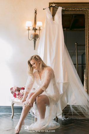 Bride in sheer robe adjusting her garter, wedding dress hanging on mirror in elegant room.