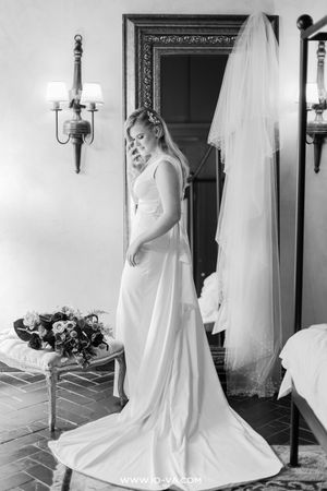 Bride in elegant white gown with veil, standing by mirror holding bouquet in black and white photo.