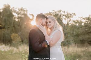 Bride and groom share a romantic golden hour embrace outdoors, with warm sunset light glowing through trees behind them.