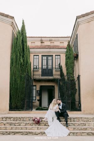 Bride and groom sitting on stone steps before a Mediterranean-style villa with tall cypress trees.