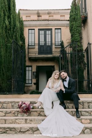 Bride and groom sitting on stone steps before an Italian-style villa, surrounded by cypress trees.