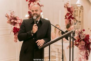 A smiling man in a black suit speaks into a microphone at an elegant wedding reception with red floral arrangements.
