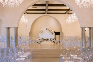 Elegant wedding ceremony venue with crystal chandeliers, ghost chairs, floral arch altar, and white arched walls.