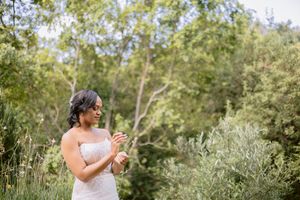 A bride in a white lace gown stands among lush green trees, smiling and holding a small bouquet outdoors.