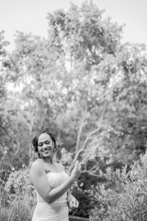 Black and white photo of a smiling bride in a strapless lace gown posing outdoors among lush trees.