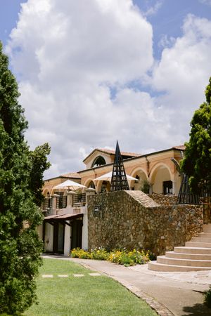 Stone villa with arched facade, pyramid sculpture, manicured gardens, and umbrella-lined terrace under a partly cloudy sky.