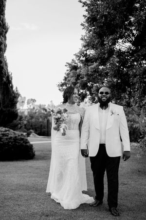 Black and white photo of a smiling bride and groom in formal wedding attire standing outdoors among lush trees.