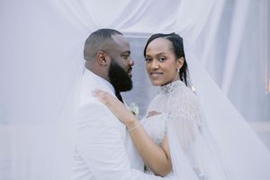 A bride and groom in white wedding attire pose romantically under a flowing white veil backdrop.
