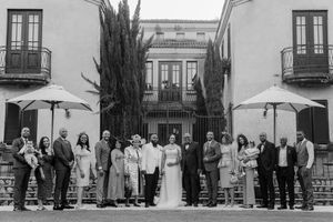 Black and white wedding party photo in front of elegant venue with cypress trees and umbrellas.