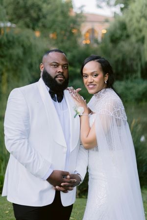 Elegant bride and groom in white wedding attire posing outdoors, showcasing their rings with lush greenery backdrop.