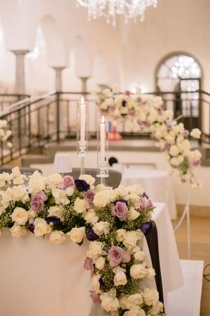 Elegant wedding reception table adorned with white and purple roses, candles, and floral arrangements in a grand venue.