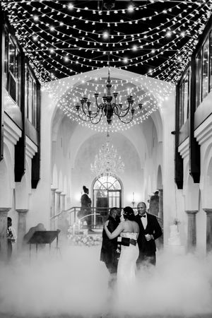 Black and white wedding photo of couple dancing under string lights and chandeliers in elegant venue with misty floor effect.