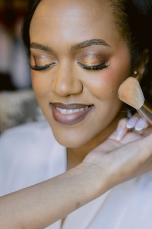 A bride with glowing makeup and false lashes gets blush applied with a fluffy brush on her wedding day.