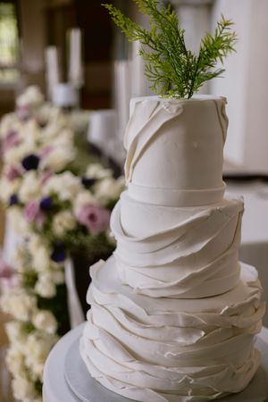 Elegant four-tier white wedding cake with ruffled fondant layers and fresh green sprig topper, displayed near floral arrangements.